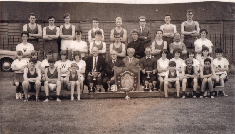 Back row, left to right: C. Gee (senior champion); A. Parry (junior champion) E. Smith, J. Morley, E. Bollard, J. Lynch, J. Wright (secretary), B. Chalton. Third row: B. Banks, E. Kendrick (youth champion), J. Watkins (senior boys' champion), R. Tarrant, D. Burns, I. Wilson (junior boys' champion), R. Hewson, A. Sheridan. 
Second row: J. Hartley. S. Elliot, V. Metcalf, M. Ashcroft, Y. Broomhead, Dr. N. Bleasdale. Mr. Aldridge, Mr. J. Edward (president), Mr. Parry, D. Hughes (senior girls' champion),  Jones, L. Tunstall, G. Houston, S. Gill (junior girls' champion). 
Front row: L. Watcham, M. Pugh, J. Bleasdale, M. Gee, D. Adair, E, Griffith.
28/07/1964 outside clubhouse in Leasowe Road (Sloar Campus) after the club track and field championships
